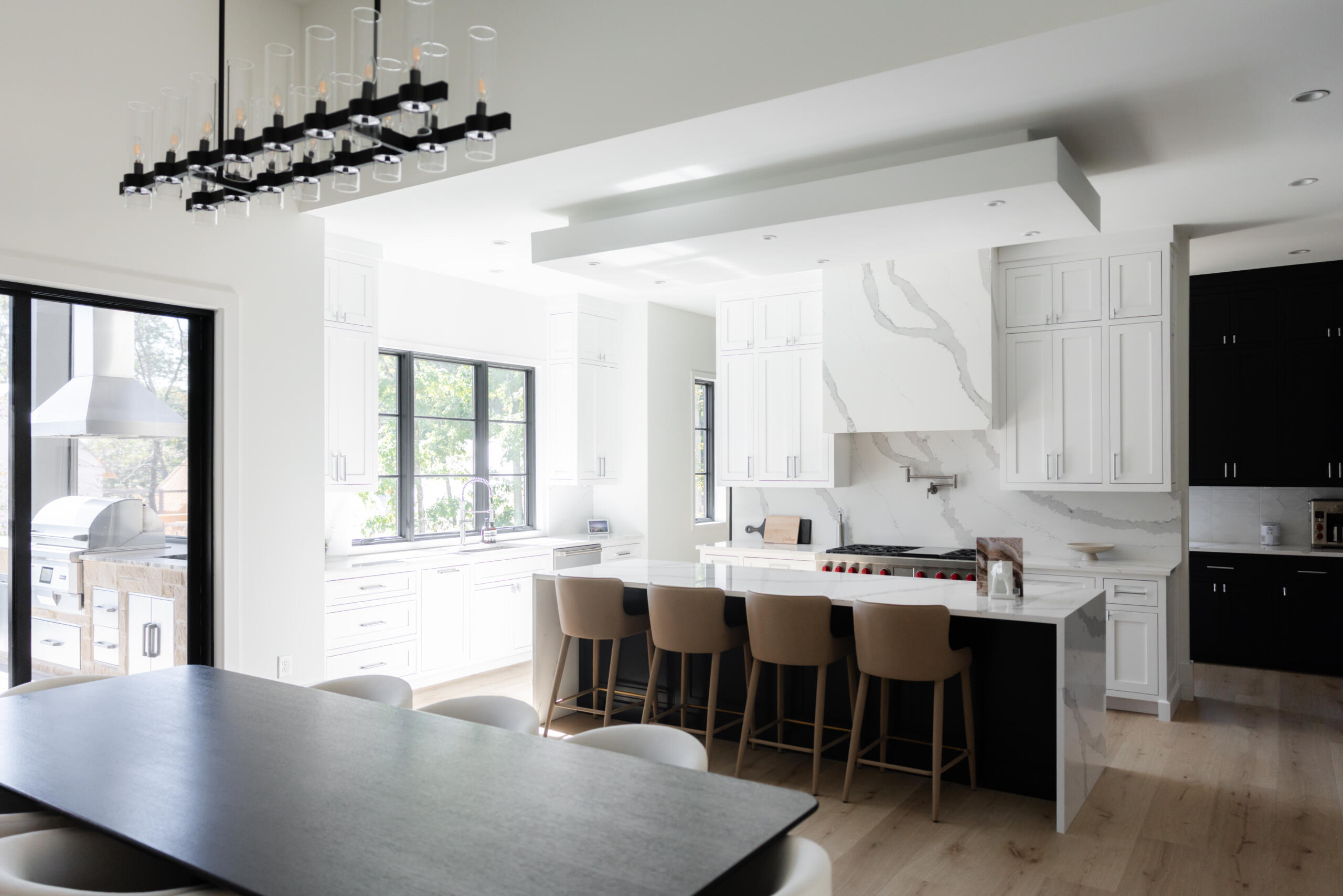Luxury kitchen with white cabinets, black linear pendant, marble island, bar stools, dark navy cabinets, and outdoor kitchen view - Southern Luxury Homes